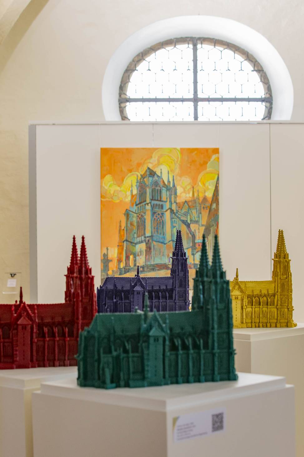 Mehrere farbige Miniaturnachbildungen des Regensburger Doms in einer Ausstellung, im Hintergrund ein Gemälde des Doms unter Wolken mit orangefarbenem Himmel.